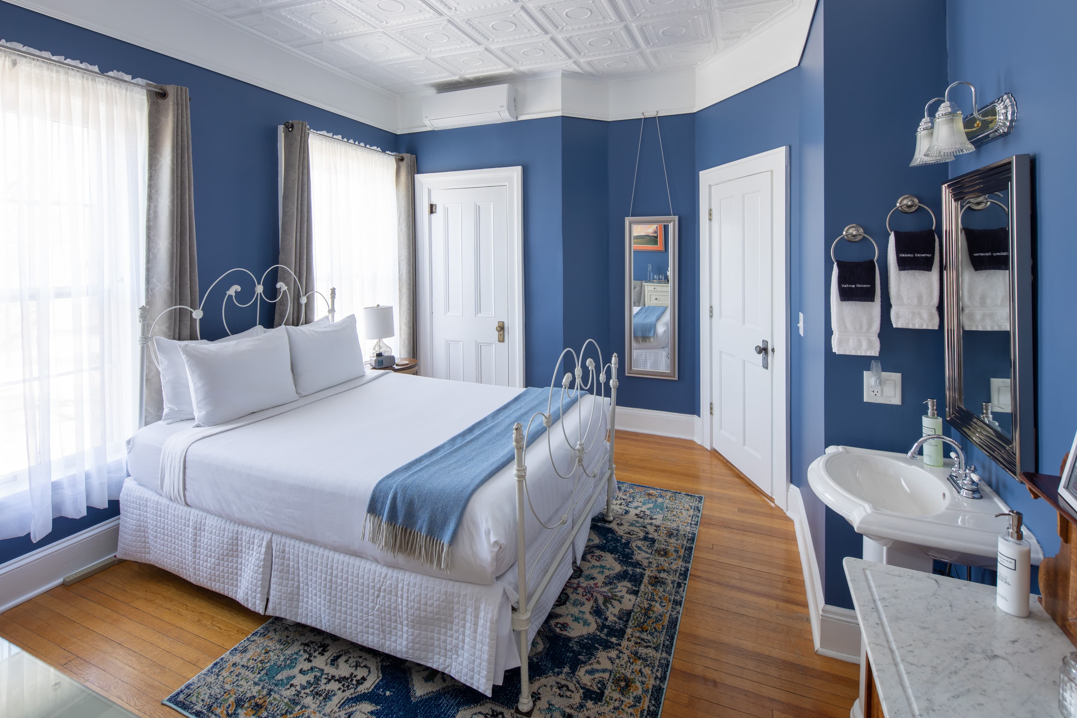 A bedroom with bold blue walls, a white king (recently upgraded) brass bed with a blue throw, and hardwood floors. A small white sink is in the corner.