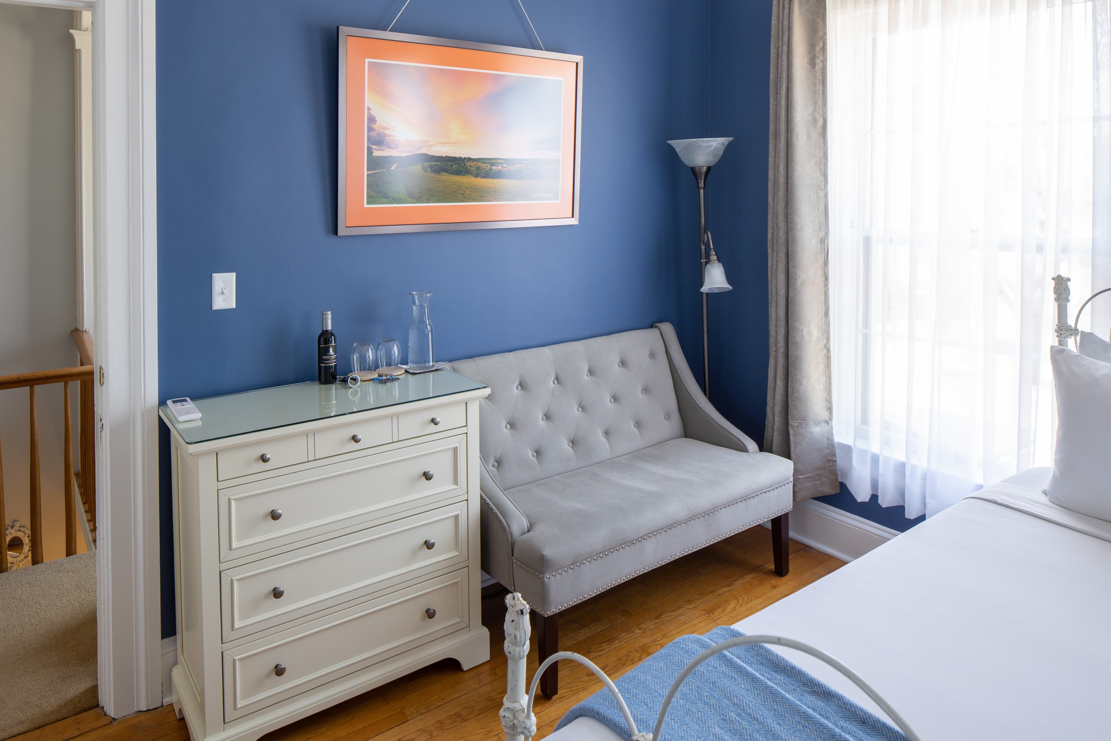 A cozy bedroom with blue walls, a light gray loveseat, and a white dresser next to a window with gray curtains.