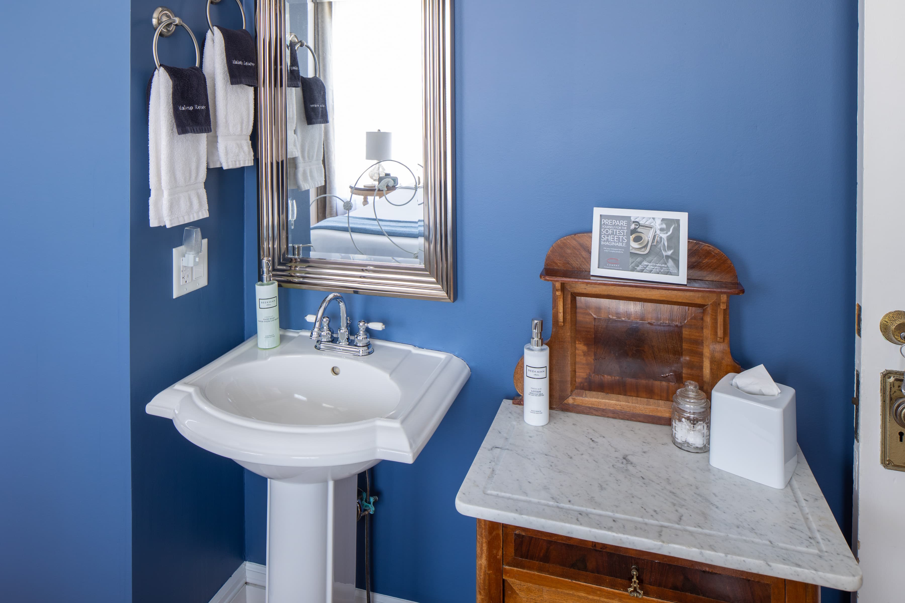 A corner of a room with a white pedestal sink against a bold blue wall. Next to it is a small, vintage wooden dresser with a marble top.