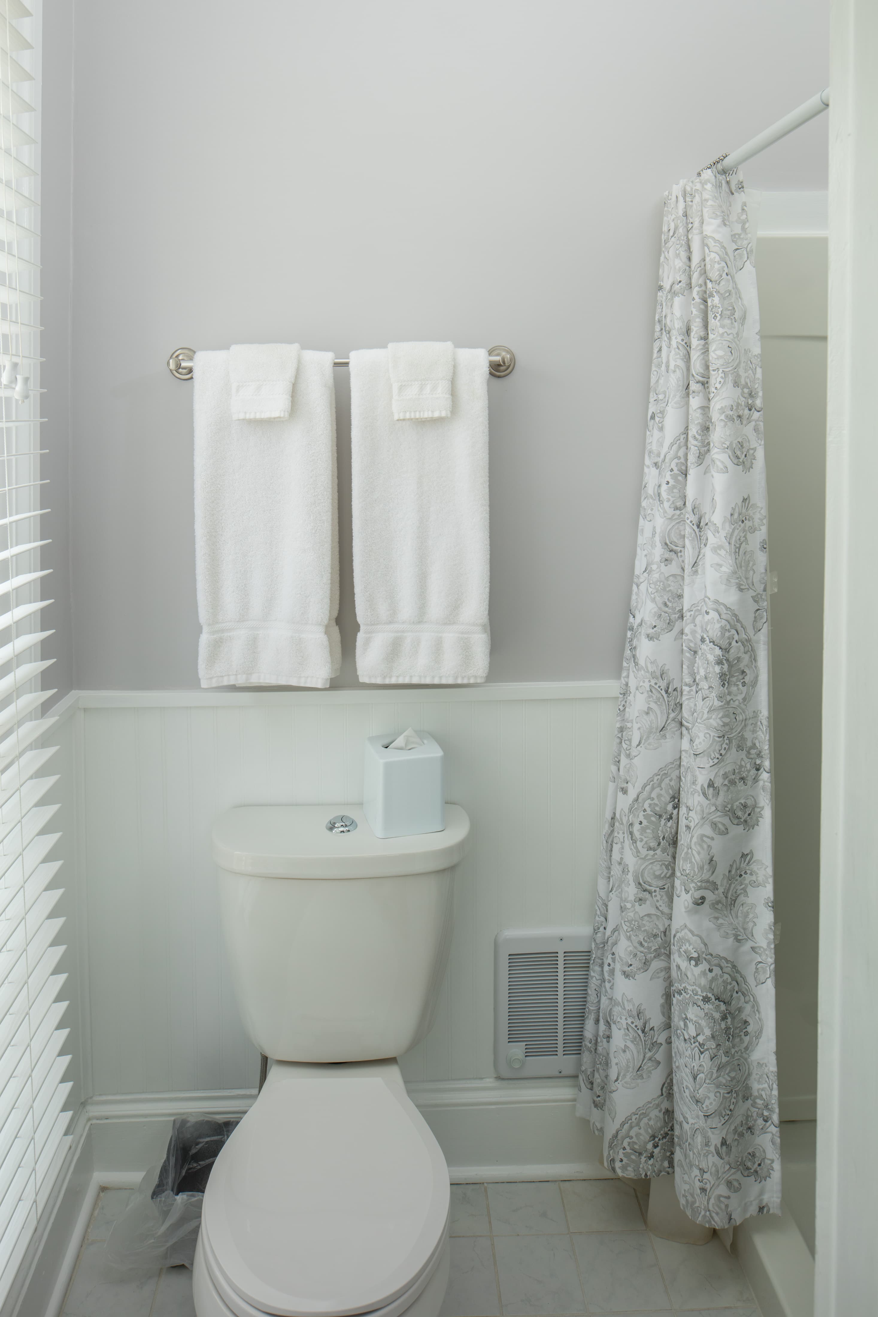 A simple bathroom with white wainscoting and light gray walls. A toilet sits in the foreground, with two white towels hanging above it. A gray and white patterned shower curtain hangs to the right.