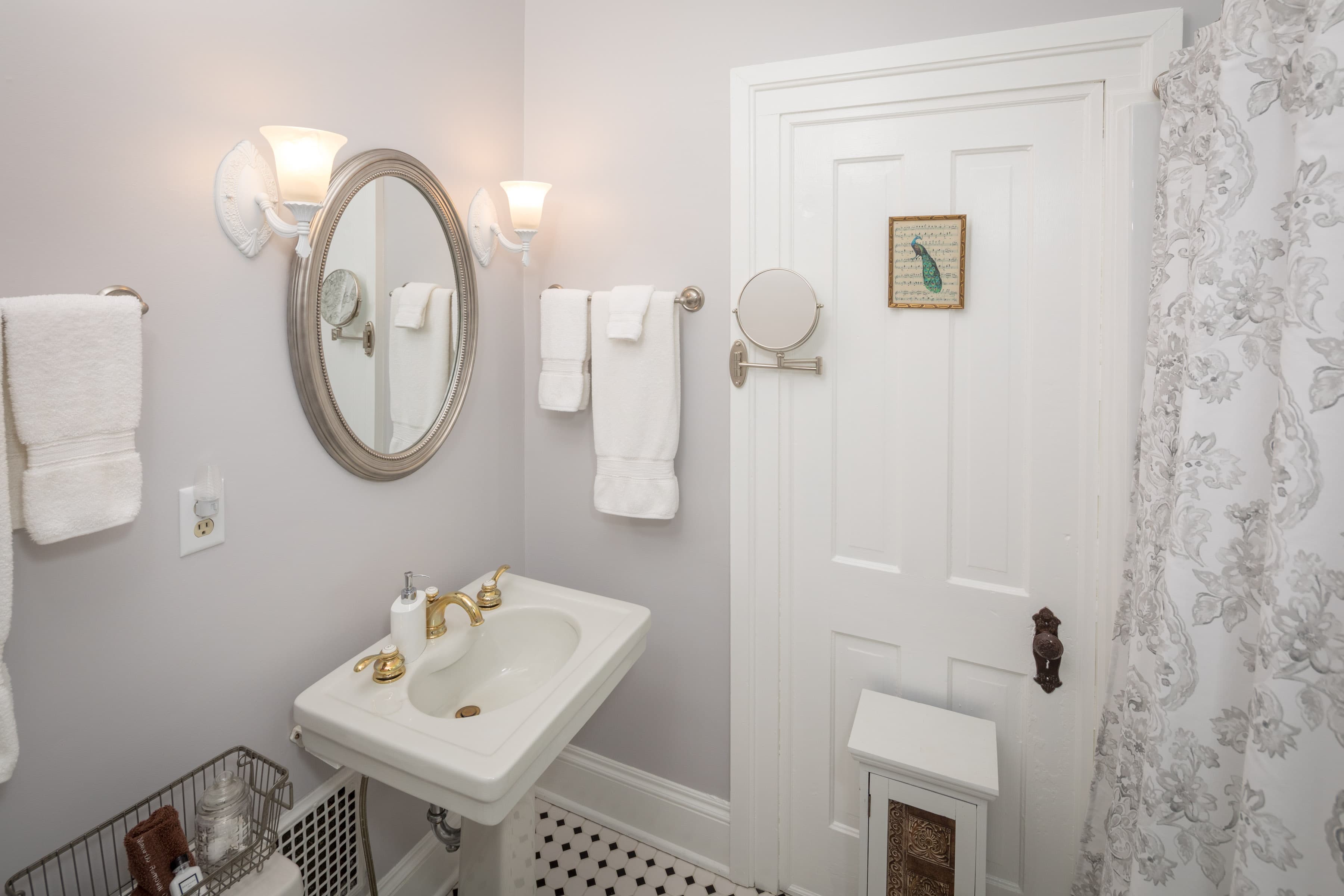 A clean, light-colored bathroom with a pedestal sink and gold fixtures. The sink is positioned below an oval mirror with a decorative silver frame. White towels are neatly folded on racks to either side of the mirror. The black and white patterned tile on the floor adds a touch of visual interest.