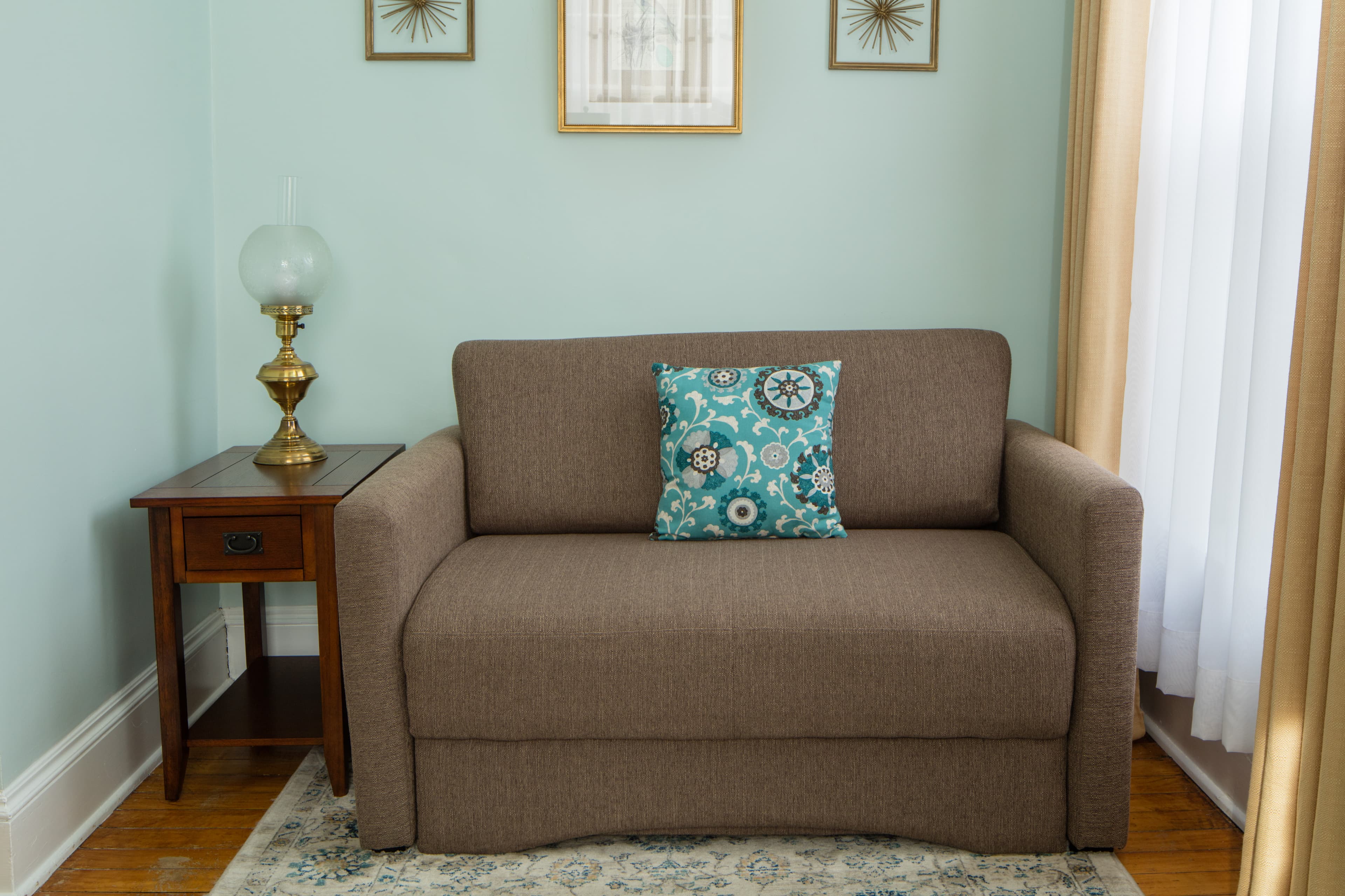 A comfortable brown sofa bed with a patterned blue and white pillow sits in a room with forest green (recently changed) walls. A small wooden side table with a vintage lamp is to the left.