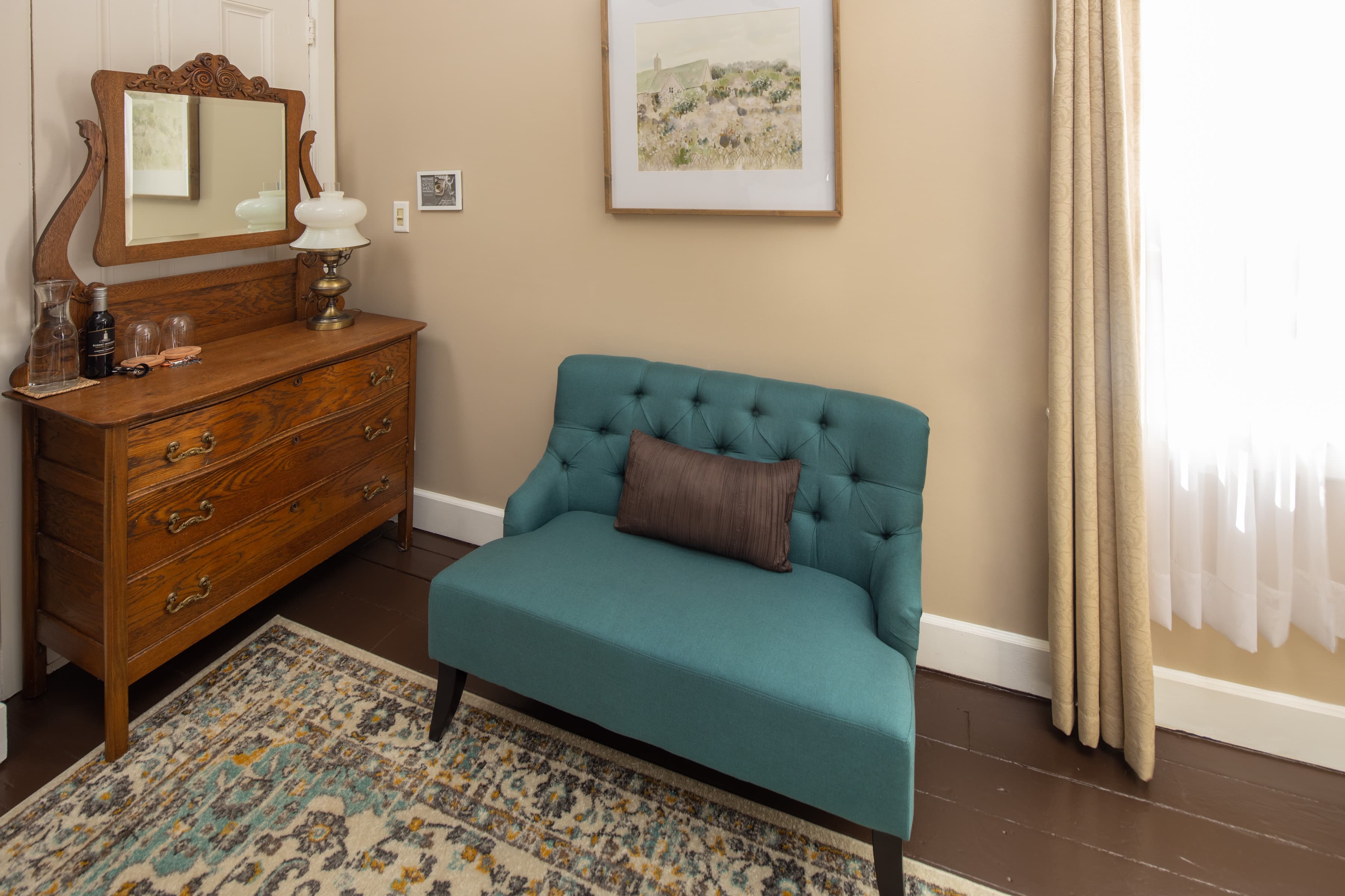 A cozy corner with light tan walls, a vintage wooden dresser, and a teal tufted chair with a brown pillow. A patterned rug is on the floor.