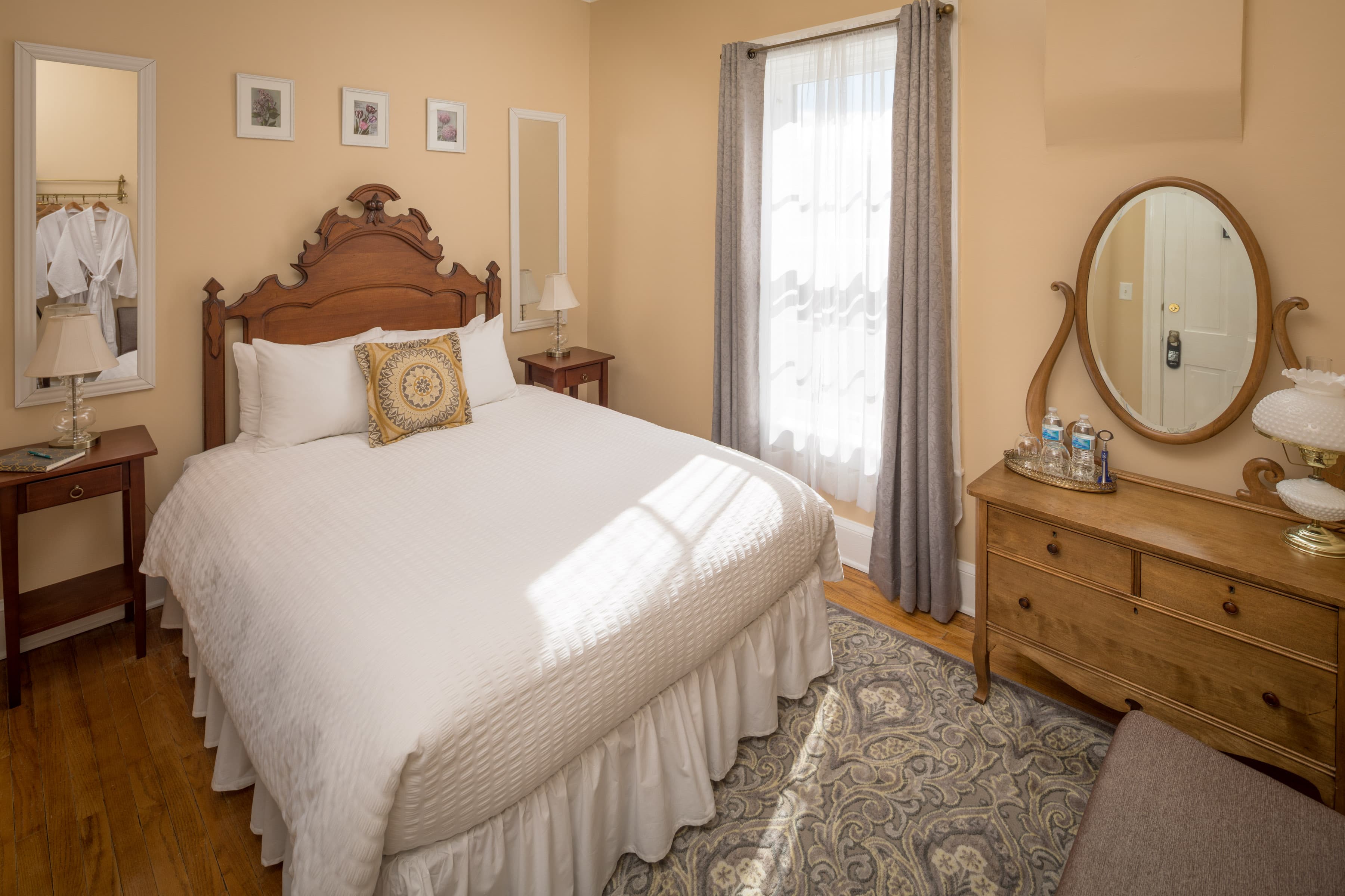 A charming bedroom with light gold walls and hardwood floors. A queen bed with a carved wooden headboard sits on a patterned rug, and a matching wooden dresser is on the right.