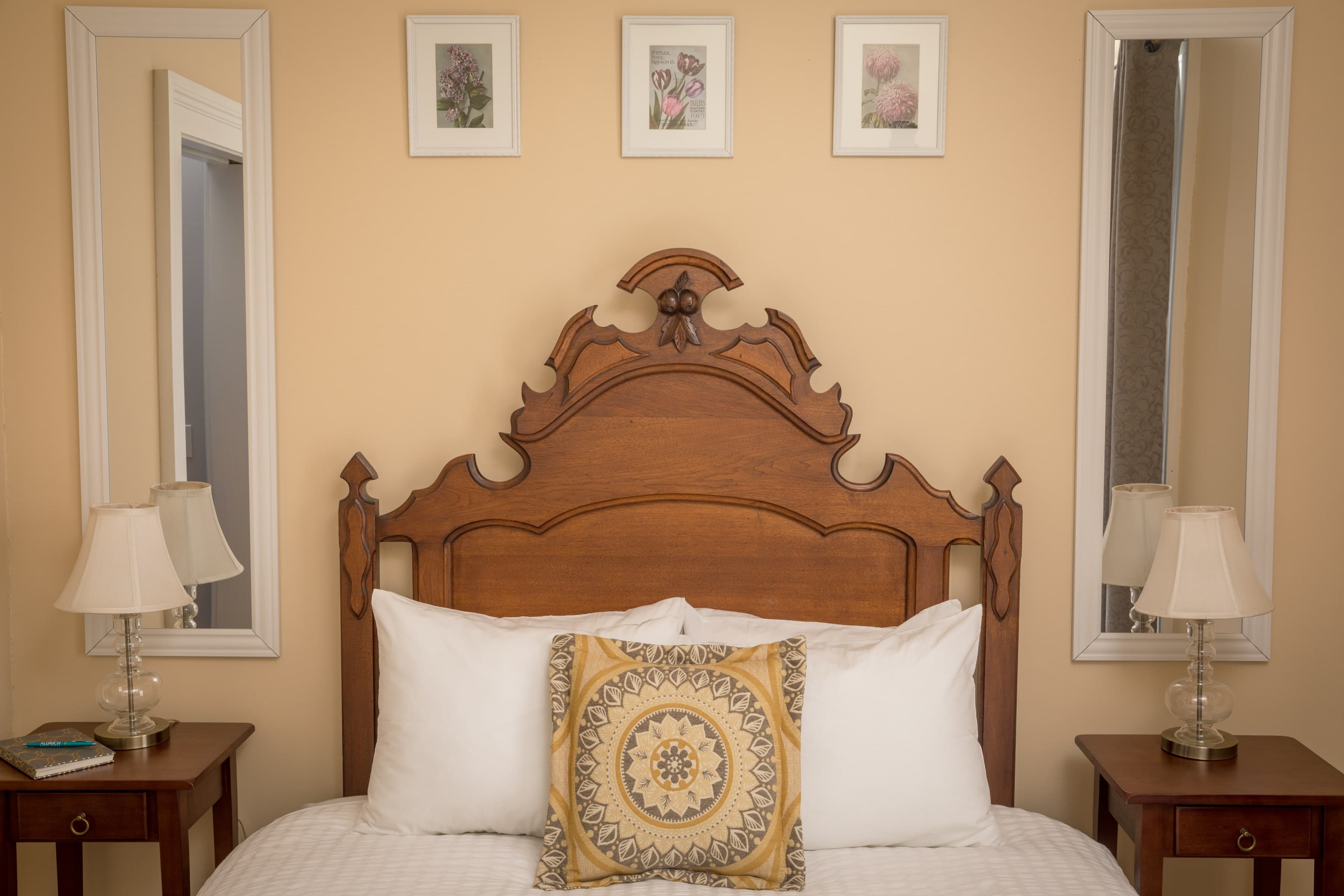 A queen bed with an antique carved wooden headboard and a decorative pillow is centered between two matching nightstands with lamps and mirrors on a light gold wall.