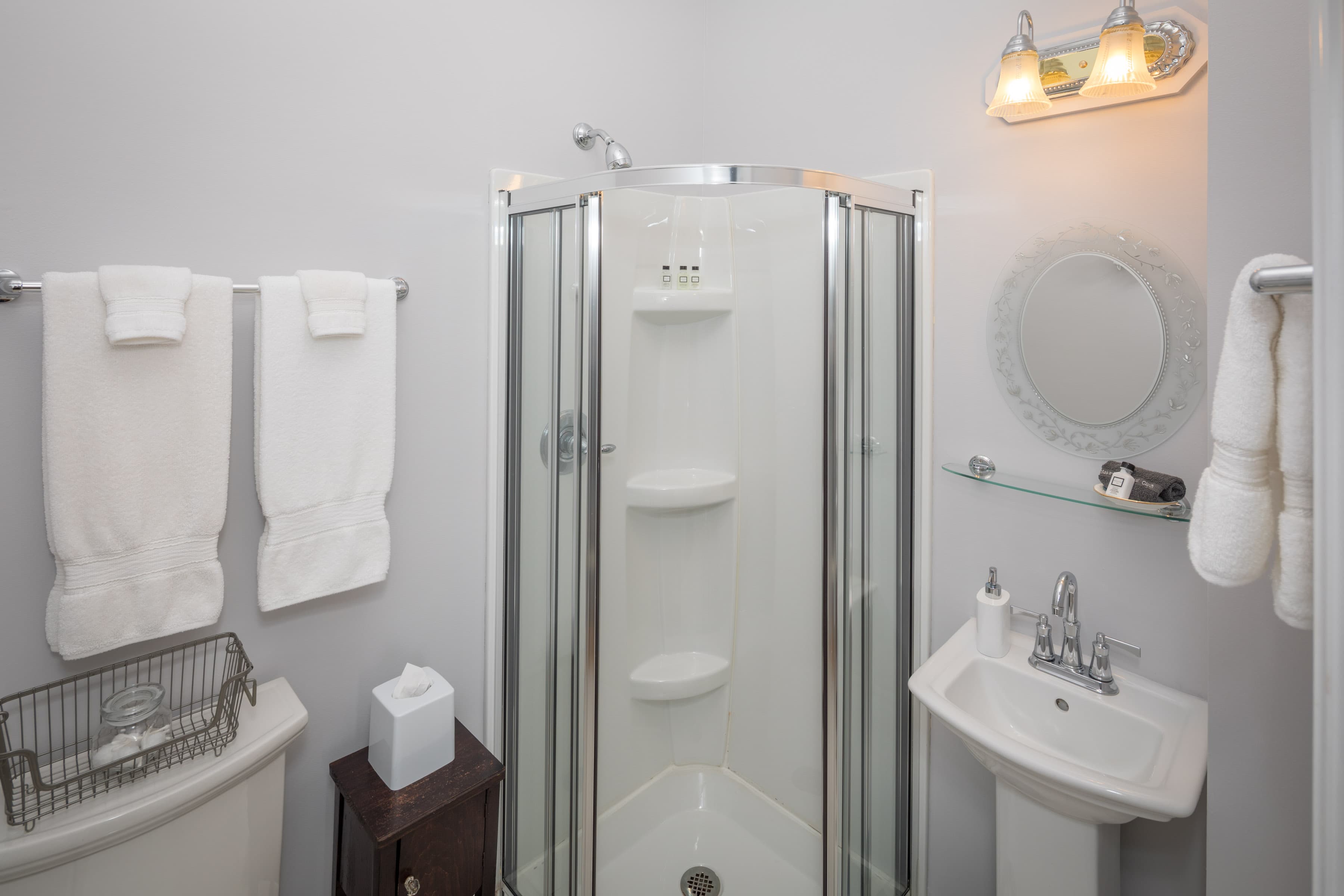 A bright bathroom with light gray walls, a glass shower stall, a pedestal sink, and a toilet. White towels are neatly folded on a rack.
