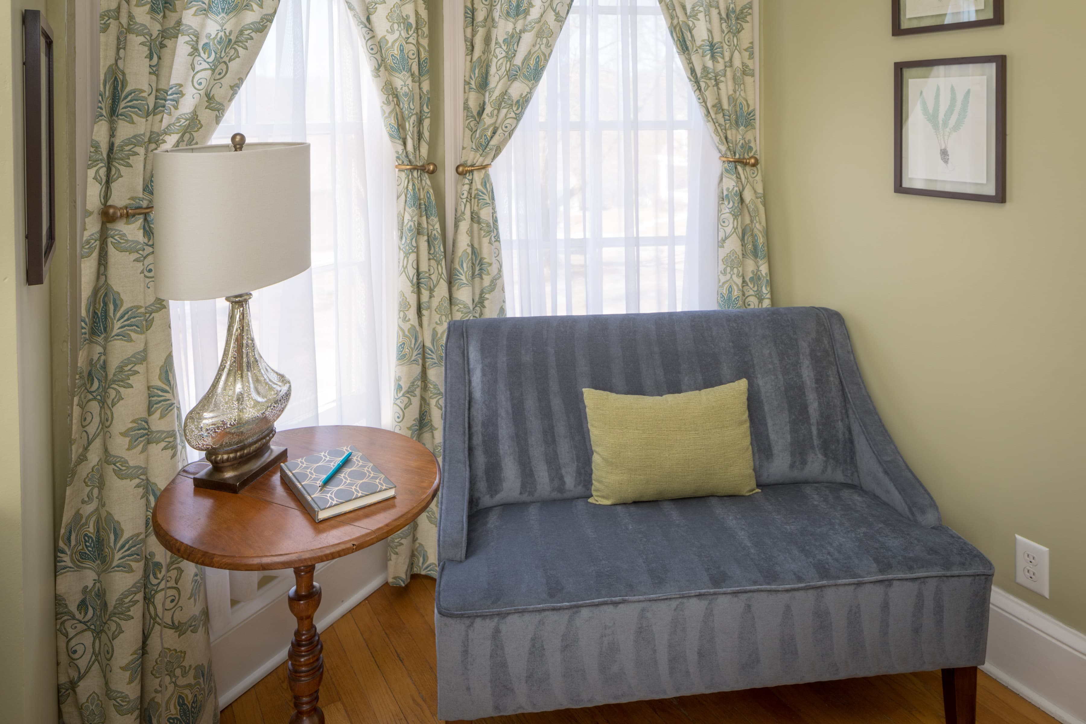 A quiet reading nook with a velvet loveseat and a small wooden side table with a lamp. A small, light green pillow rests on the loveseat.