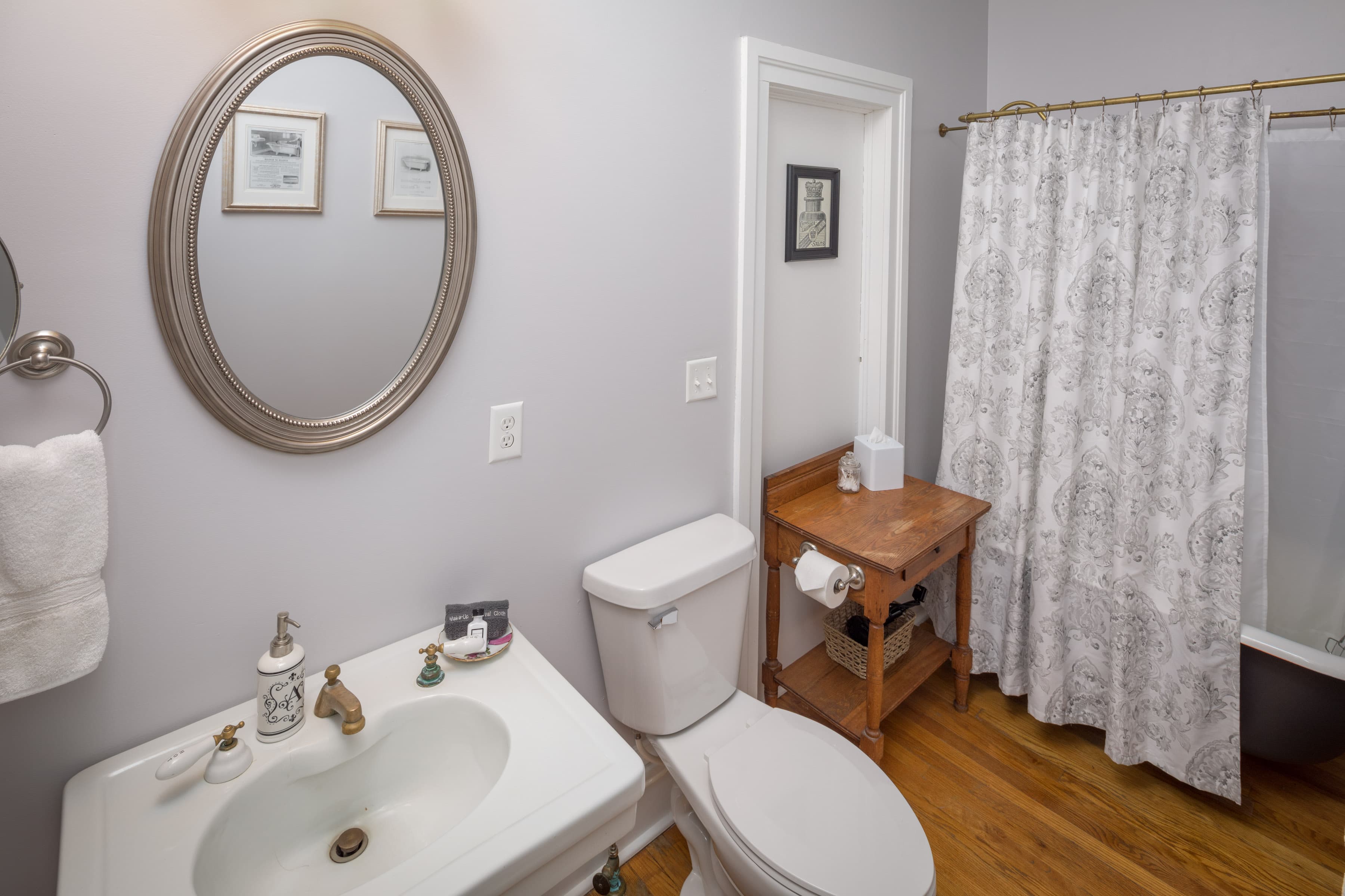A cozy bathroom with light gray walls, a white pedestal sink with a mirror above it, and a toilet. A patterned shower curtain hangs over a clawfoot tub/shower (this will be upgraded this winter 2026 with a luxury walk-in shower with multiple showerheads and a bench).
