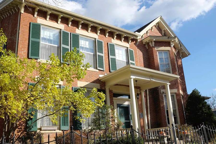 A brick house with green shutters and a front porch, surrounded by trees and a fence.