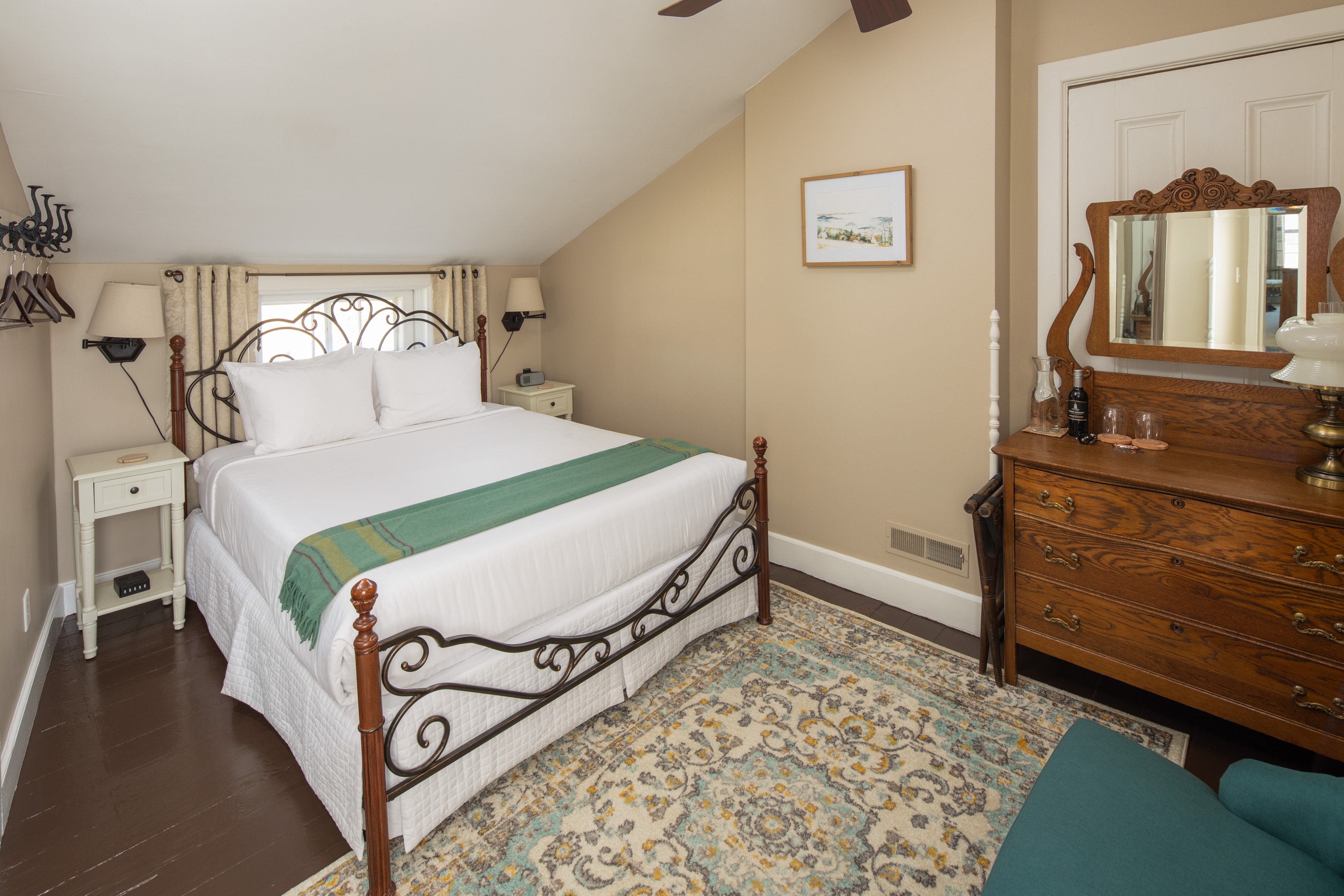 A cozy bedroom with a sloped ceiling and wrought-iron bed with a white comforter and green blanket, sitting on a patterned rug. A vintage dresser with a large mirror is on the right.