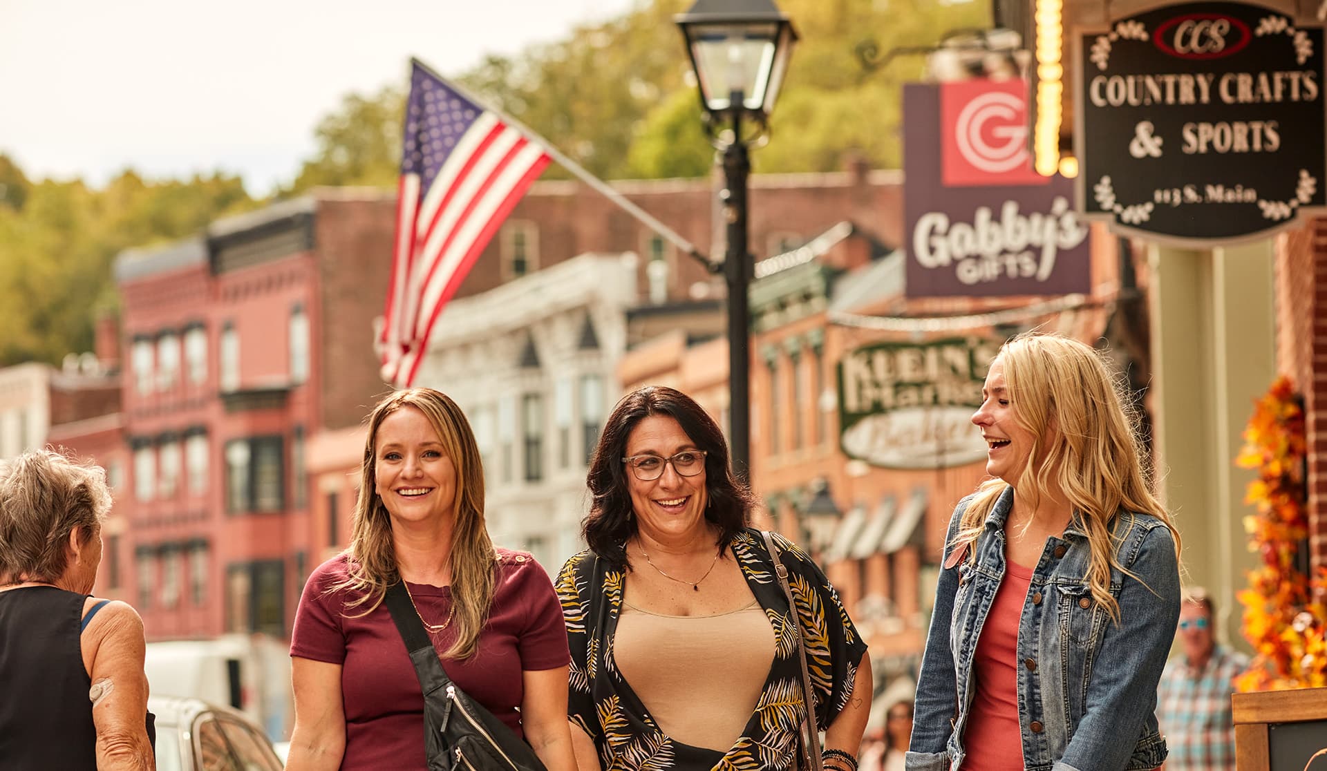 Three smiling women walk together on a vibrant street with storefronts and an American flag in the background.