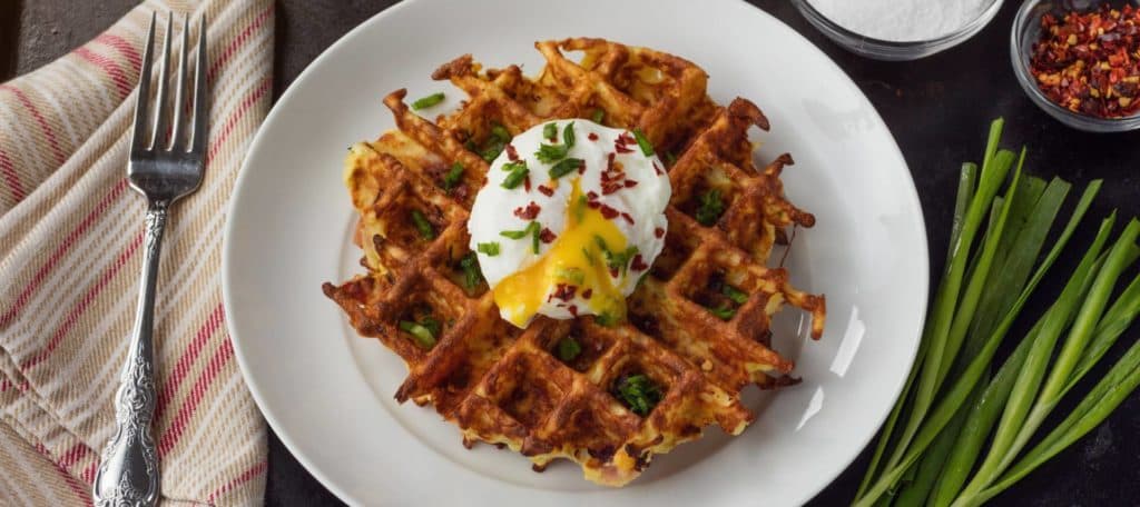A golden waffle topped with a poached egg and sprinkled with chives and chili flakes, served on a white plate.