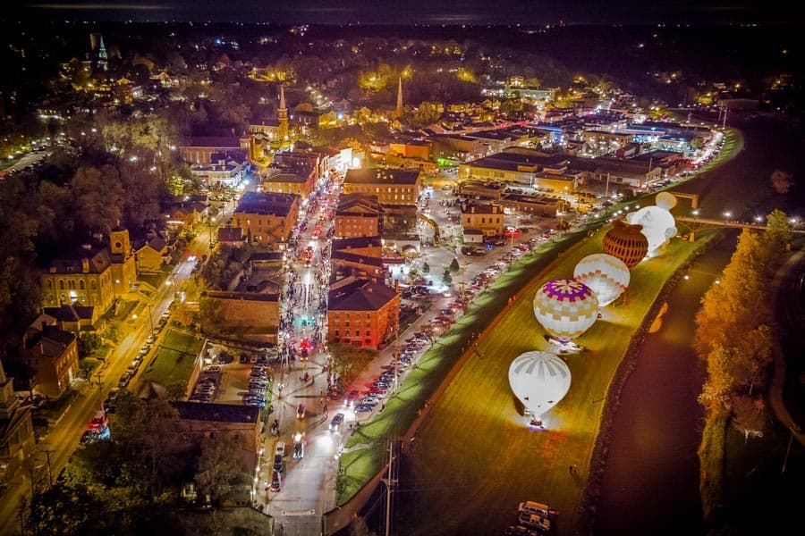 Aerial view of a vibrant, illuminated town at night with colorful hot air balloons along the riverbank.