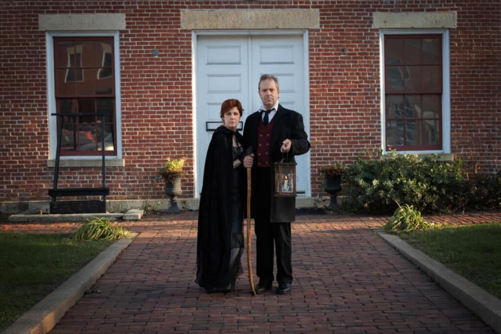 A man and a woman dressed in period clothing stand together holding a lantern in front of a brick building.