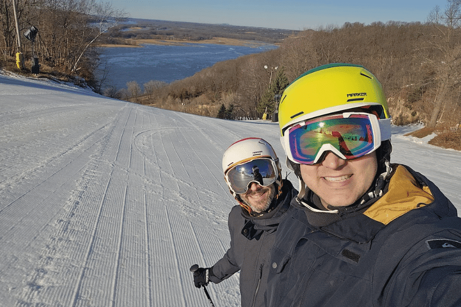 Two skiers smiling on a snowy slope with a scenic view in the background.