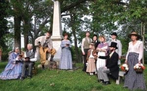 A group of people dressed in 19th-century attire gathered around a memorial in a park.