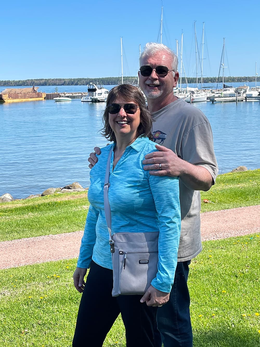 A smiling couple poses together by a marina on a sunny day.