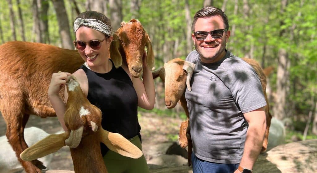 A woman and a man smile while holding goats in a wooded area.