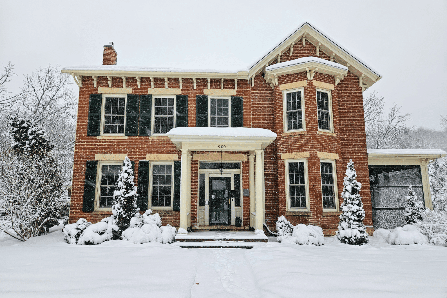 A brick house with green shutters surrounded by snow-covered trees on a winter day.