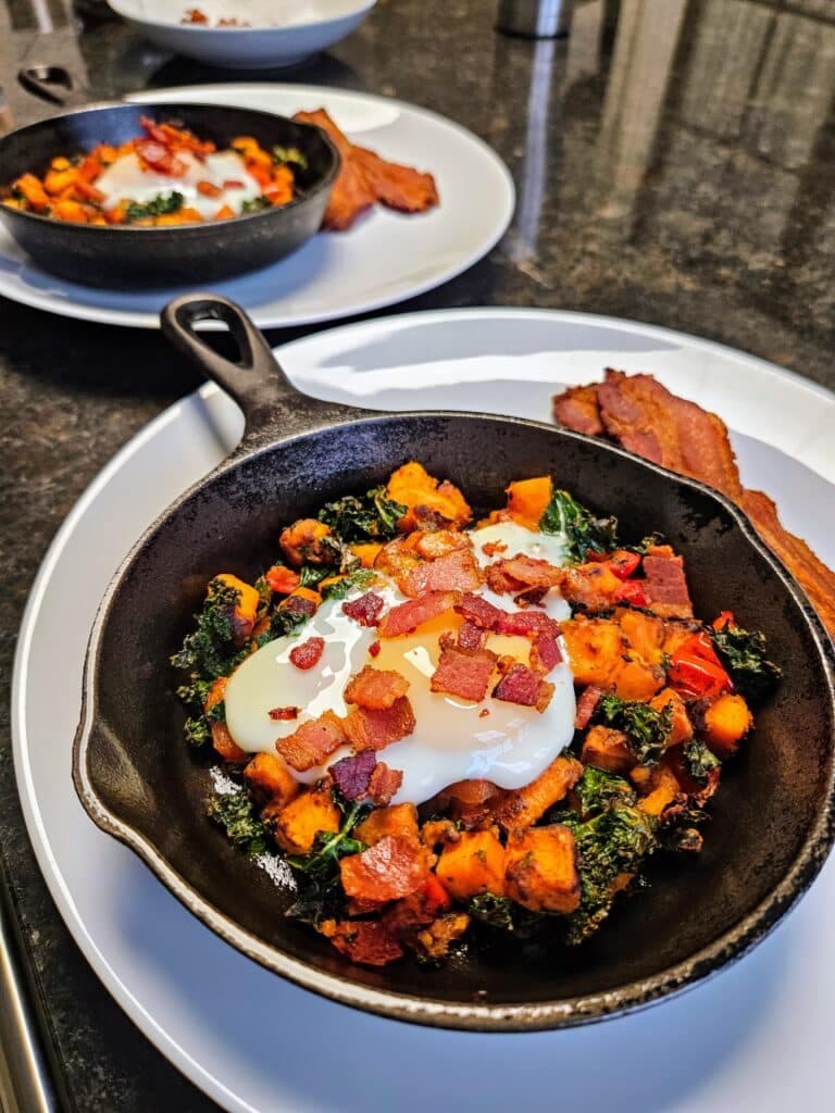 A cast iron skillet filled with sautéed kale, sweet potatoes, and topped with a poached egg and crumbled bacon.