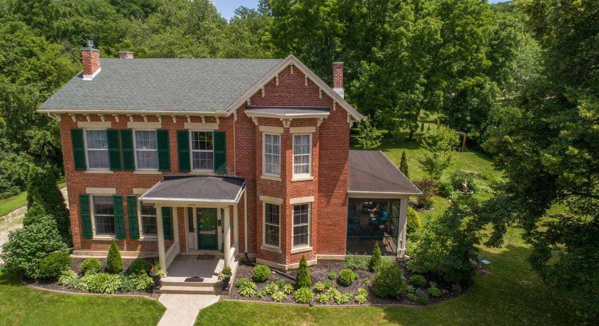 A picturesque red brick house with green shutters surrounded by lush landscaping.