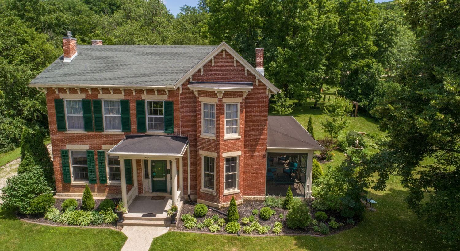 A picturesque red brick house with green shutters surrounded by lush landscaping.