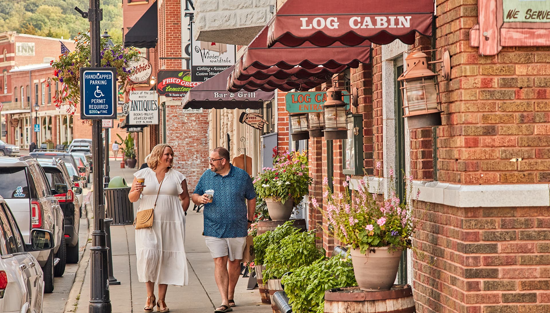 A man and woman walk arm-in-arm down a quaint street lined with shops and colorful flowers.