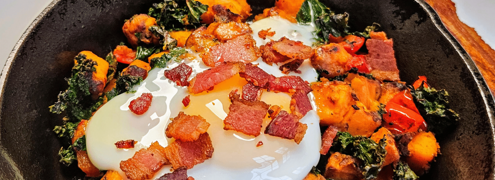A close-up of a skillet with a dish featuring a poached egg, sweet potatoes, kale, red peppers, and crispy bacon. A close-up of a skillet with a dish featuring a poached egg, sweet potatoes, kale, red peppers, and crispy bacon.