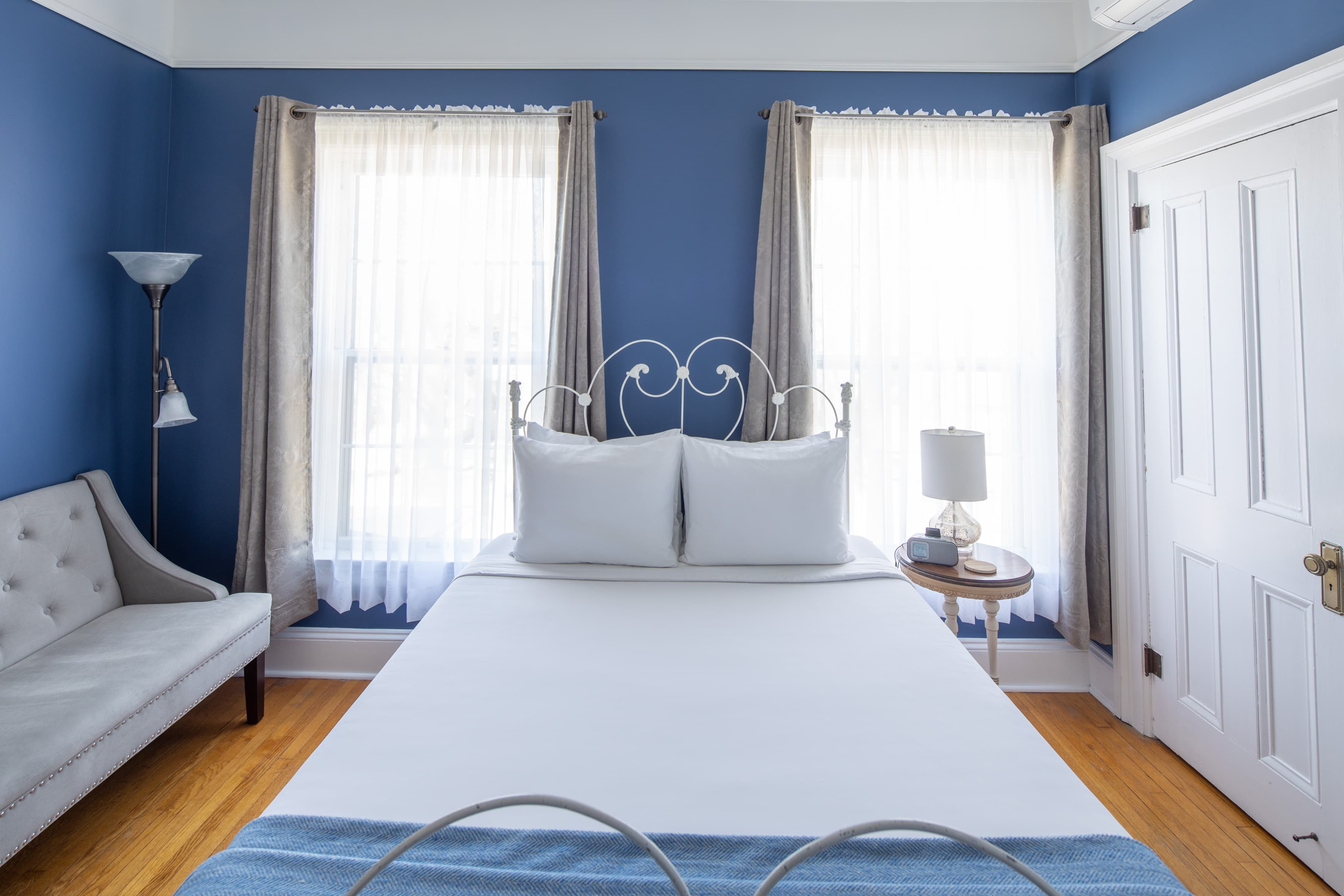 A cozy, pristine bedroom with blue walls and hardwood floors. A white king (recently upgraded) bed with a luxury brass frame sits between two large windows.