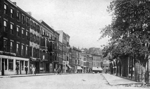 Black and white image of a historical street lined with brick buildings and shops.