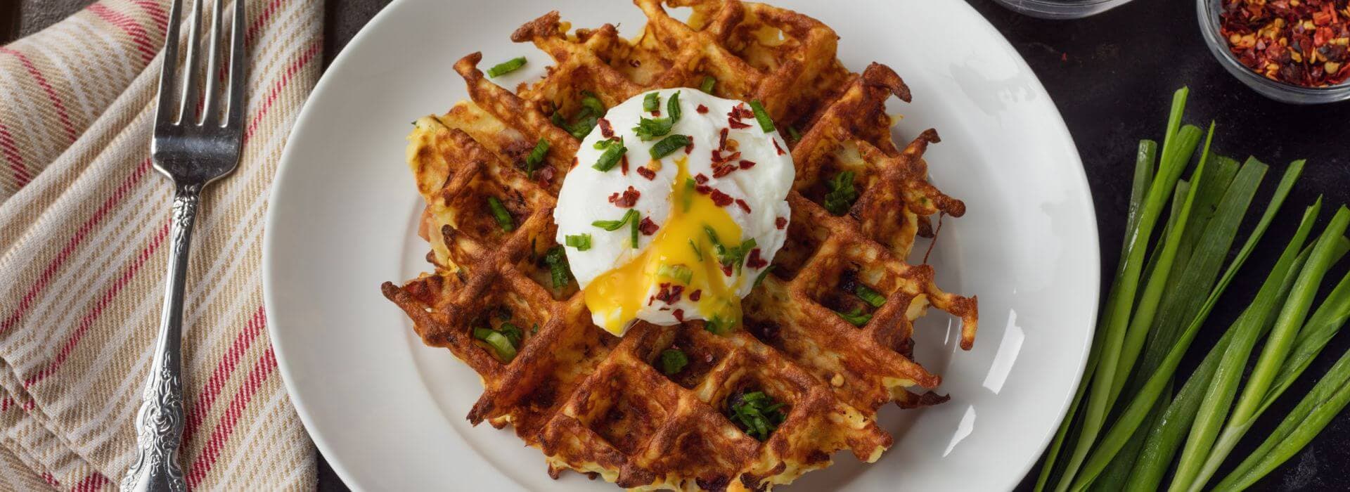 A crispy waffle topped with a poached egg, green onions, and red pepper flakes on a white plate.