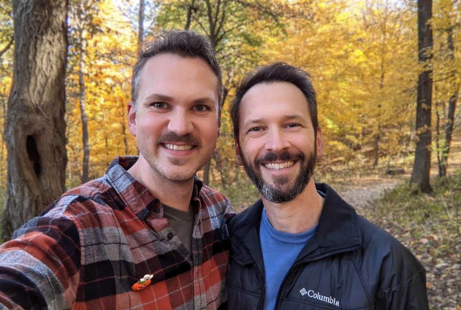 Two men smiling together in a forest with autumn foliage.