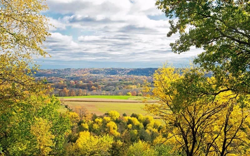 A scenic view of rolling hills and fields adorned with autumn foliage under a cloudy sky.