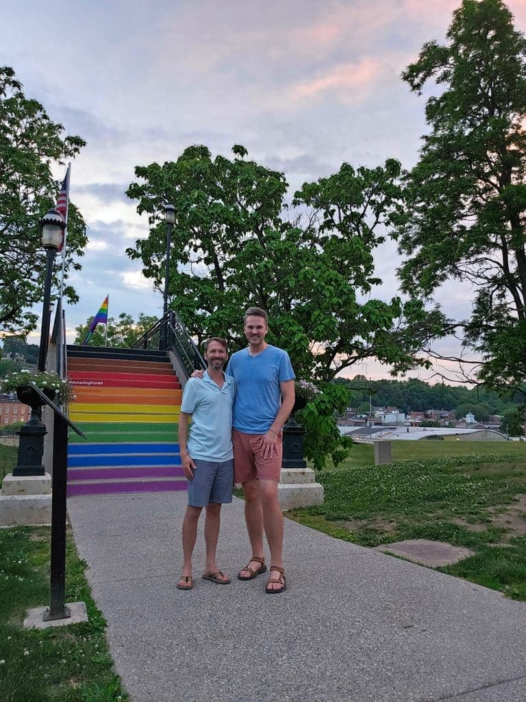 Two men stand together on a pathway beside a colorful rainbow staircase under a cloudy sky.