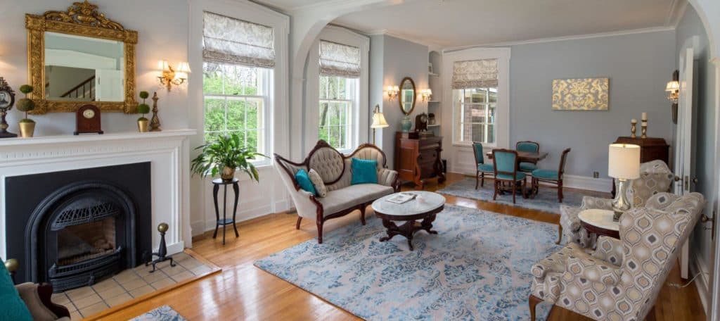 A bright, elegantly furnished living room featuring a fireplace, vintage decor, and large windows.