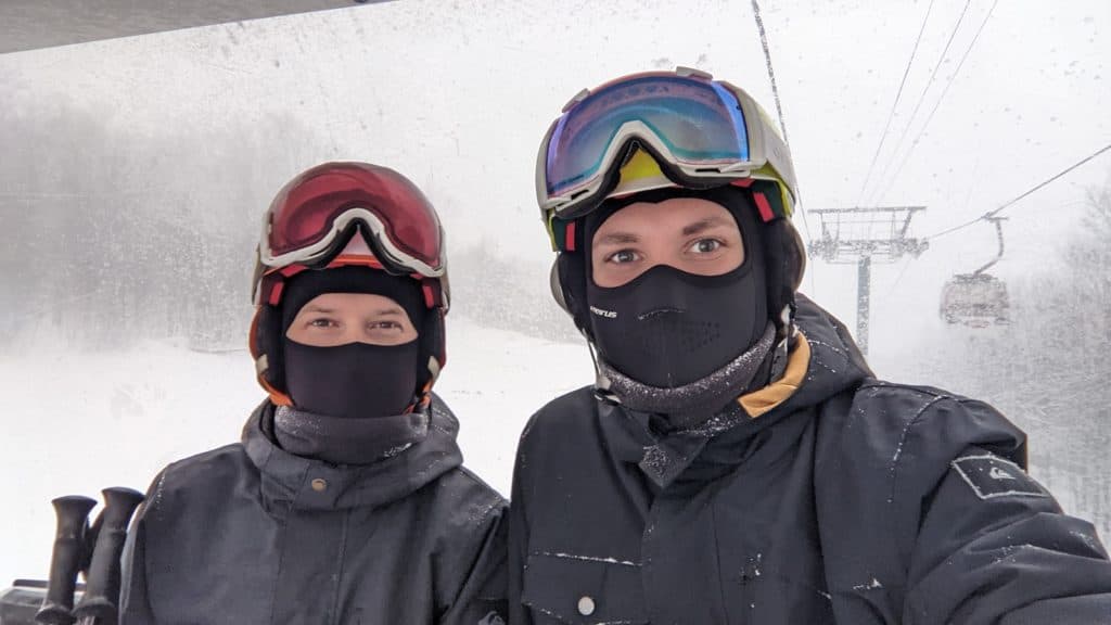 Two skiers wearing masks and goggles take a selfie on a snow-covered ski lift.
