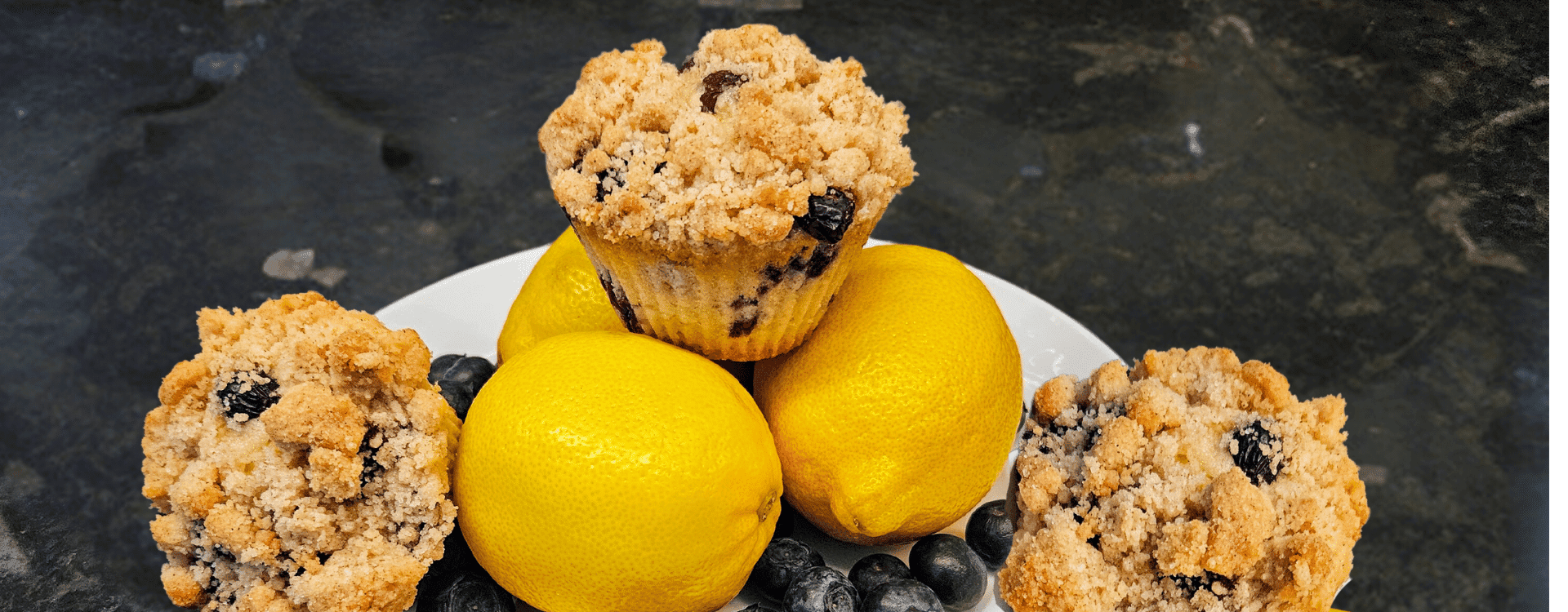 A plate holds two lemon fruits and two blueberry muffins with crumb topping.