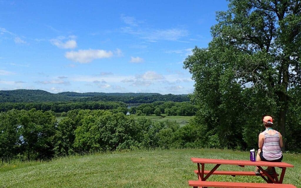A person sits on a red bench overlooking a lush green landscape and river under a blue sky.