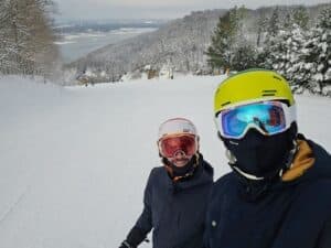 Two skiers pose for a selfie on a snowy slope surrounded by trees.