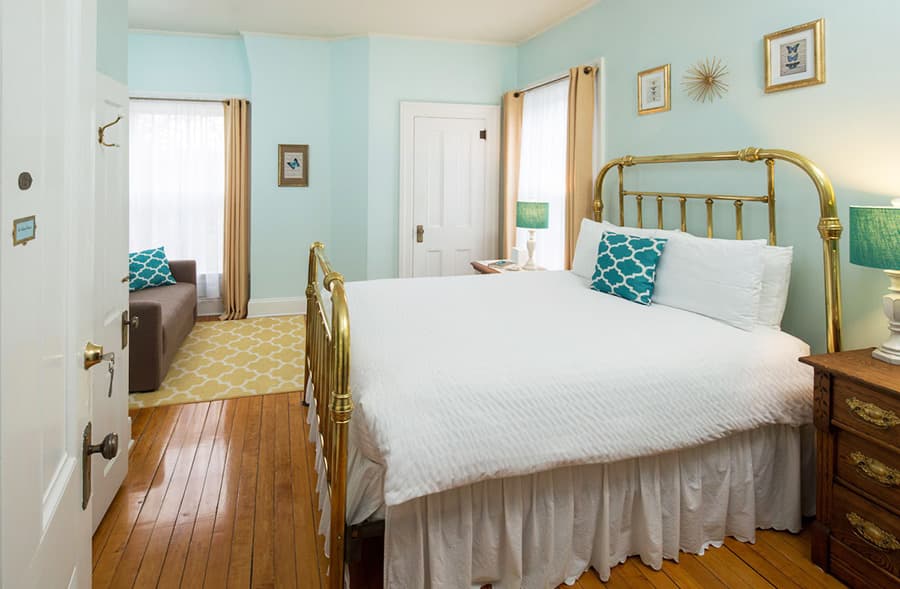 A cozy bedroom featuring a brass bed, light blue walls, and a small sitting area with natural light.