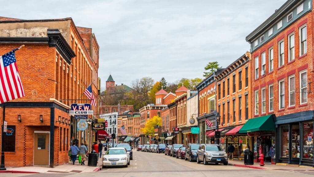A charming street lined with historic brick buildings and American flags, featuring shops and parked cars under a cloudy sky.
