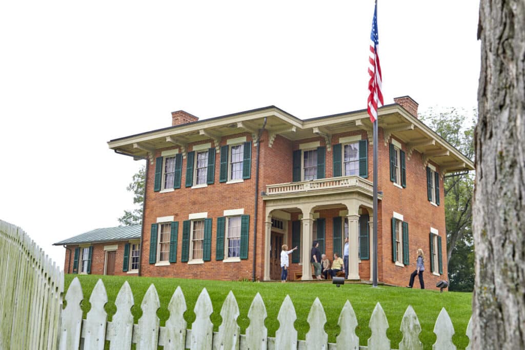 A historic brick house with a white picket fence and an American flag outside, where several people are gathered on the porch and lawn.