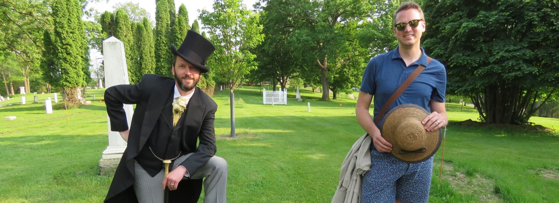 Two men, one dressed in a vintage suit and top hat and the other in casual attire, pose in a grassy cemetery.