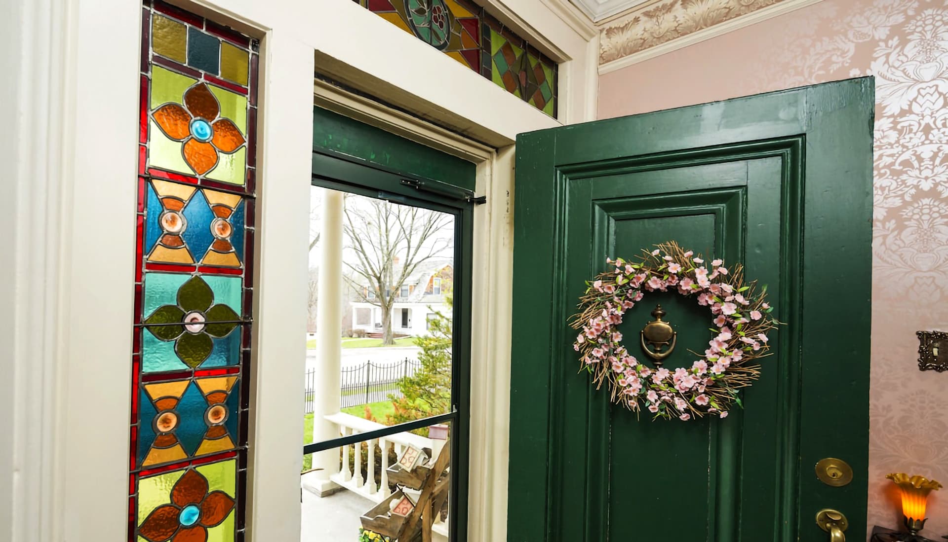A green door partially open, adorned with a floral wreath, showcasing colorful stained glass and a view of an outdoor entrance.