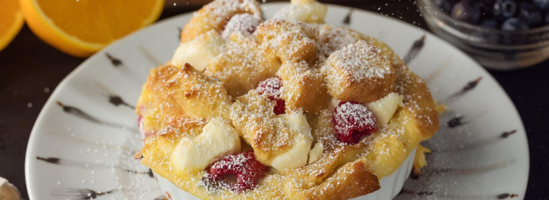 A baked dessert topped with powdered sugar, featuring bread, fruit, and a scattered background of oranges and blueberries. A baked dessert topped with powdered sugar, featuring bread, fruit, and a scattered background of oranges and blueberries.