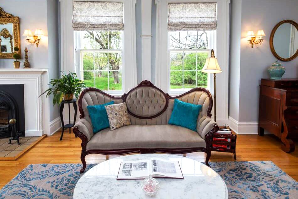 A cozy living room features a vintage upholstered sofa, large windows, fresh greenery, and elegant decor.