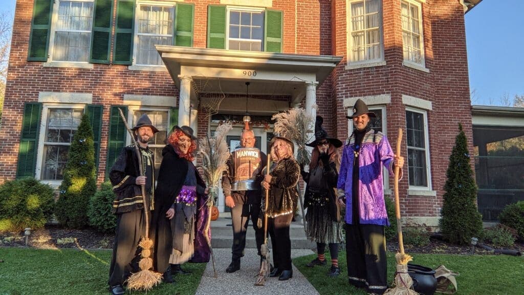 A group of six people dressed in Halloween costumes poses in front of a red brick house.