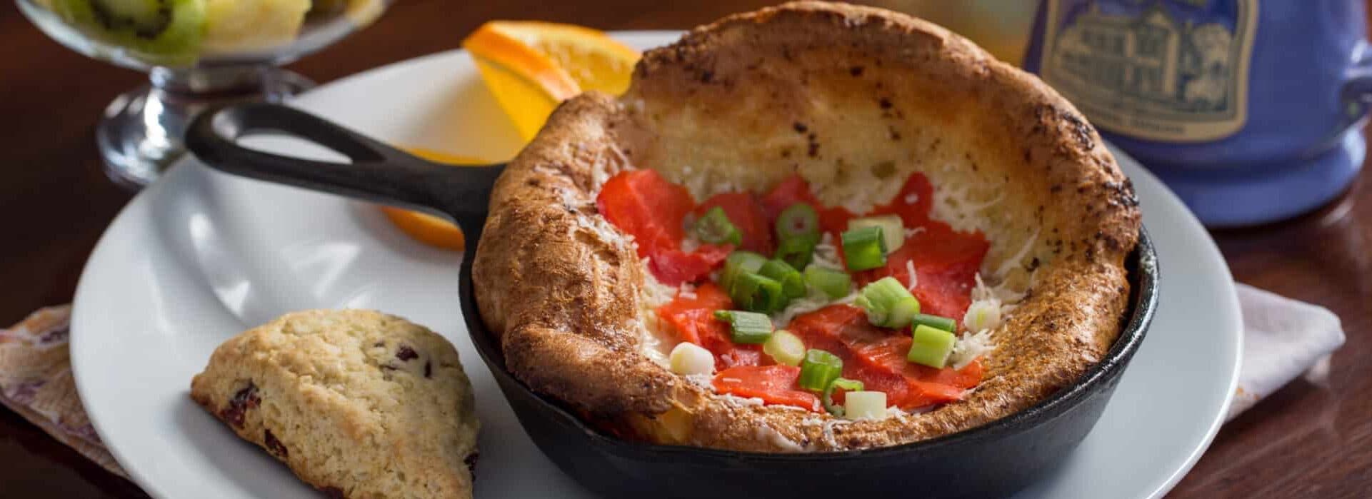 A fluffy egg dish with tomatoes and green onions served in a cast iron skillet, accompanied by a scone and orange slices. A fluffy egg dish with tomatoes and green onions served in a cast iron skillet, accompanied by a scone and orange slices.
