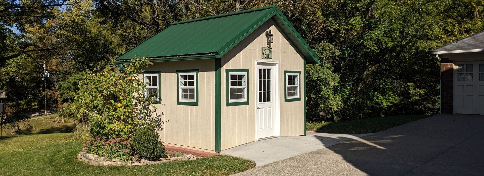 A small potting shed with a green metal roof and white walls, surrounded by greenery.