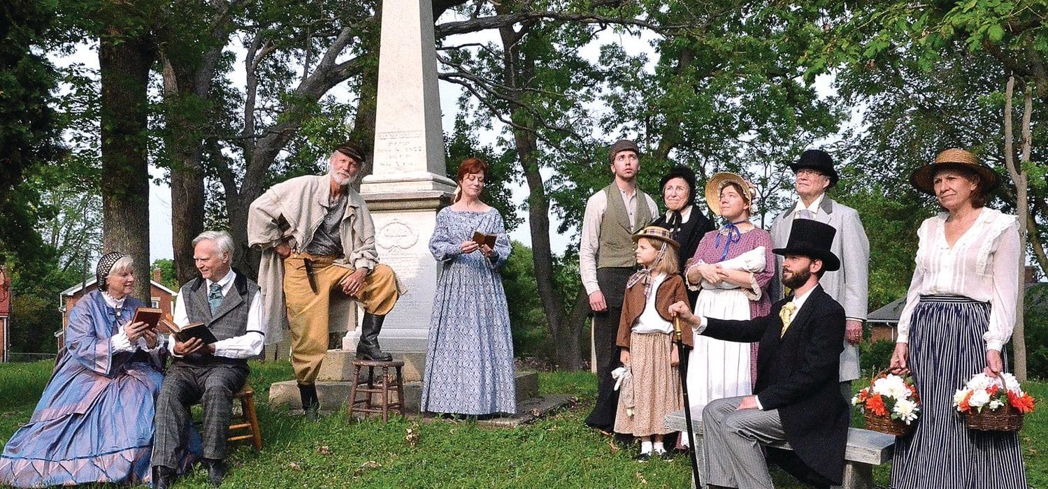 A group of people in a cemetery in front of an obelisk monument who are all dressed in 1850s attire.
